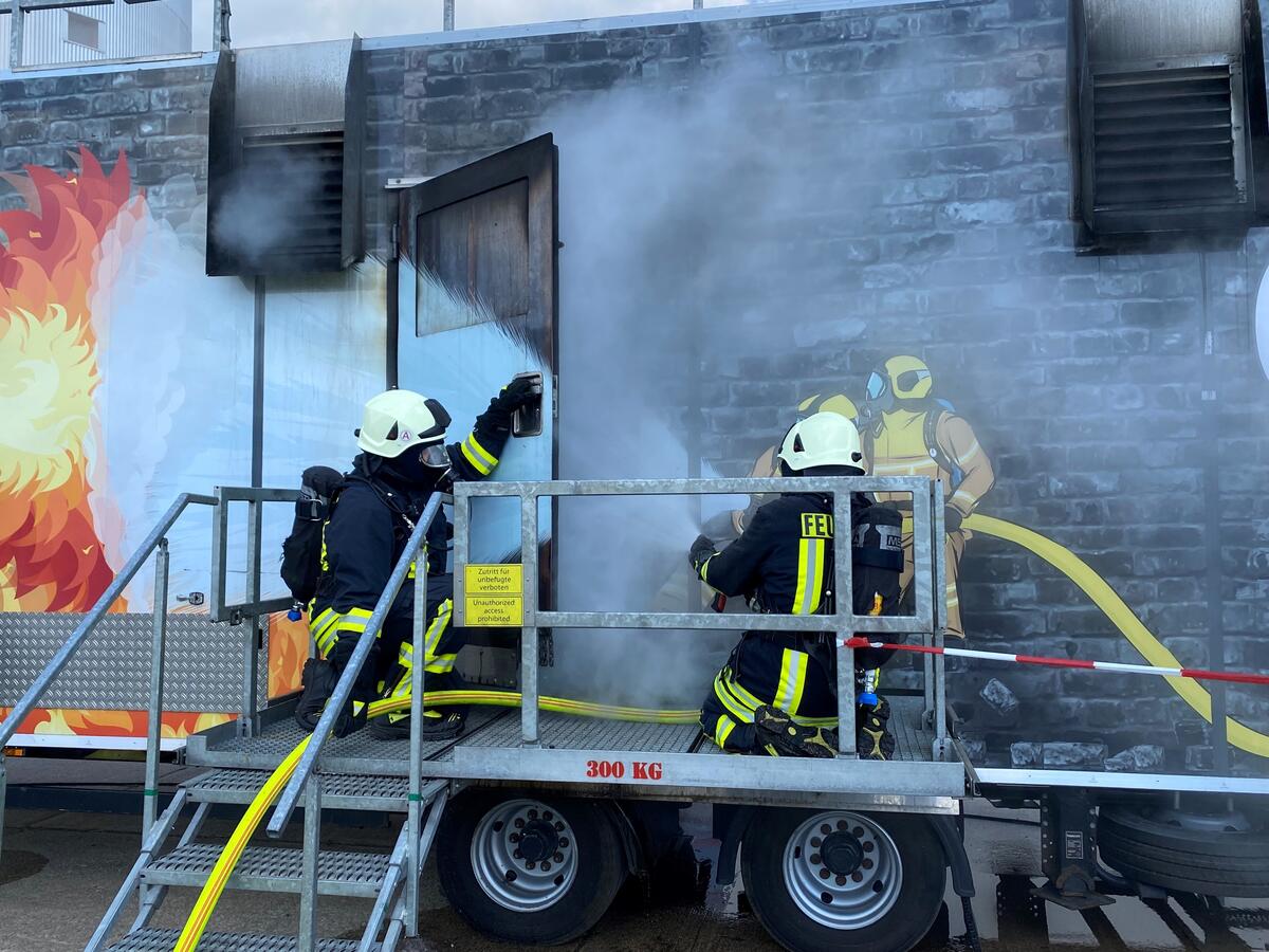 Bild vergrößern: Zwei Feuerwehrmänner öffnen die rauchende Tür des Brandübungscontainers und löschen mit Wasser.